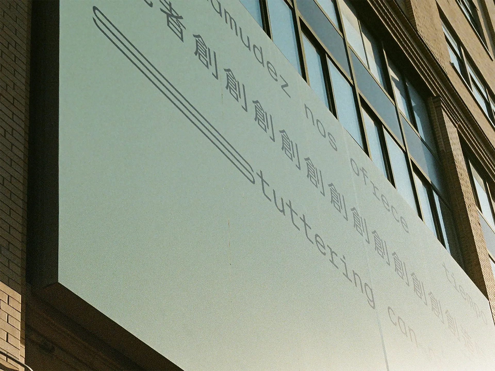 A close-up photograph of sun shining on a billboard. Three lines of black text appear on a light seafoam green background, in Spanish, Chinese and English, translating to: ‘Stuttering can create time.’ The text is in a sans serif typeface organised in three straight lines within the top half of the composition. The bottom half of the composition is empty. The text is stretched and repeated to represent stammering.